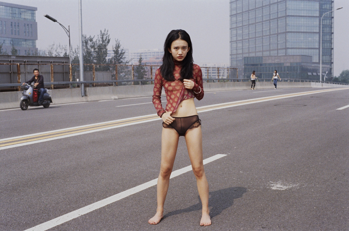 Foto av ung kinesisk kvinne midt på en bilvei iført truse og en genser hun drar opp og viser deler av magen.